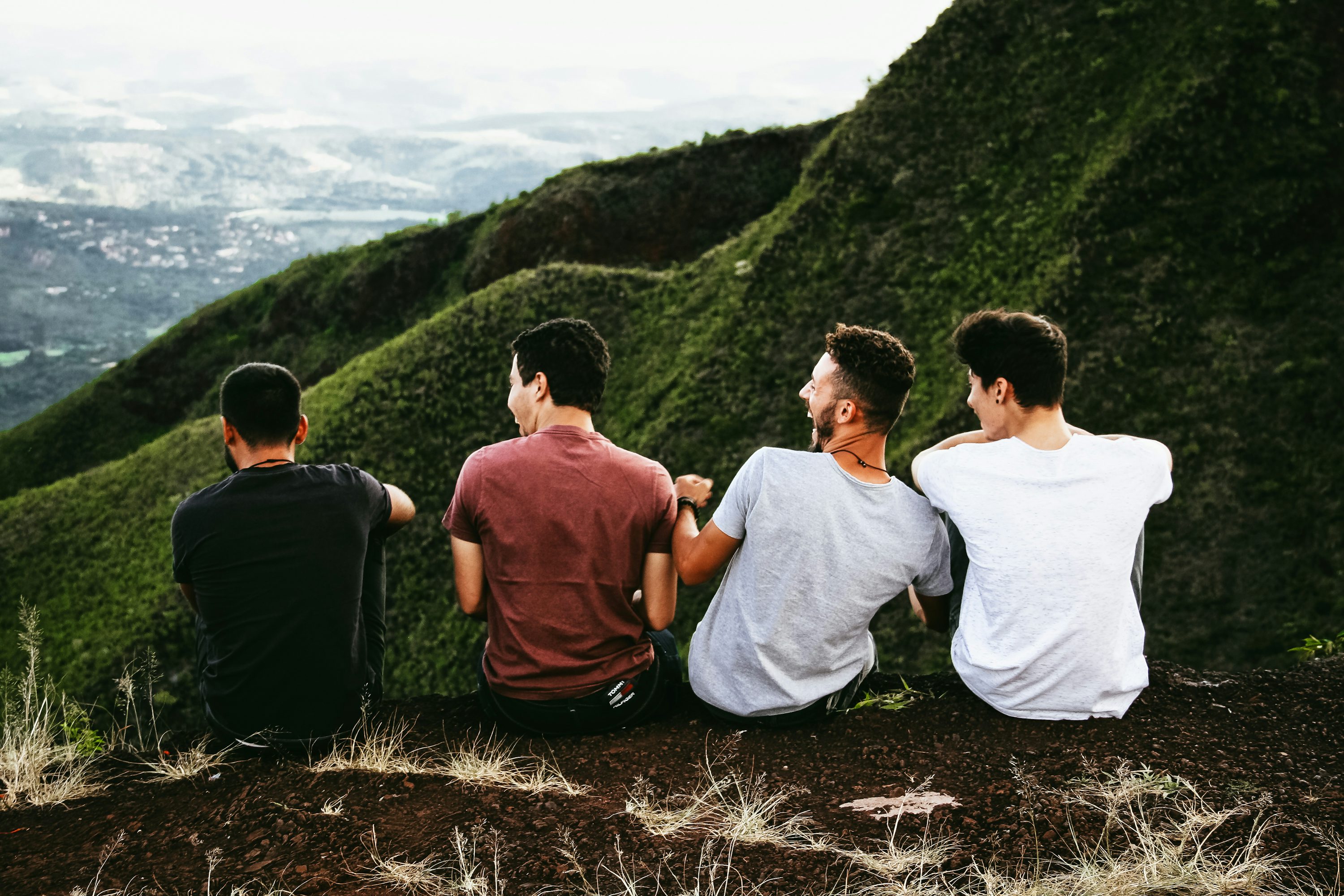 Four young men sitting together on grassy hill with scenic sky and mountains in background